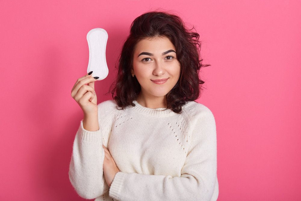 First period box for teens: Woman in white sweater holding a sanitary pad against a pink background, symbolizing menstruation or feminine hygiene.