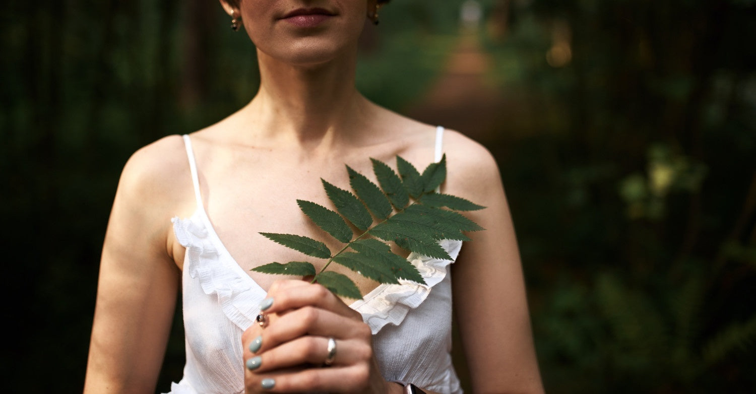 A woman in a white dress holds a green fern leaf, standing in a lush forest with soft lighting.