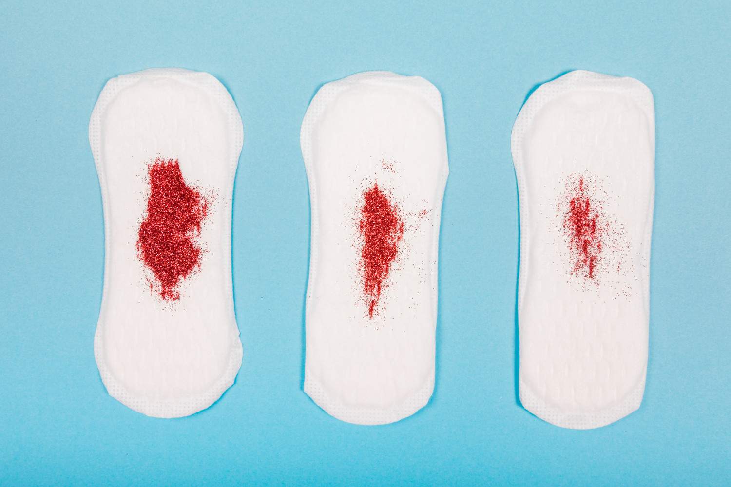 Medium flow period pads: Three sanitary pads on a light blue background, each with varying amounts of red glitter on them, simulating menstrual flow.