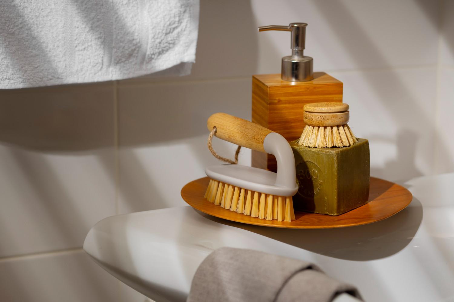 Eco-friendly bathroom swaps: A wooden soap dish holds a brush and a bar of soap, alongside a soap dispenser, with a towel hanging in the background.