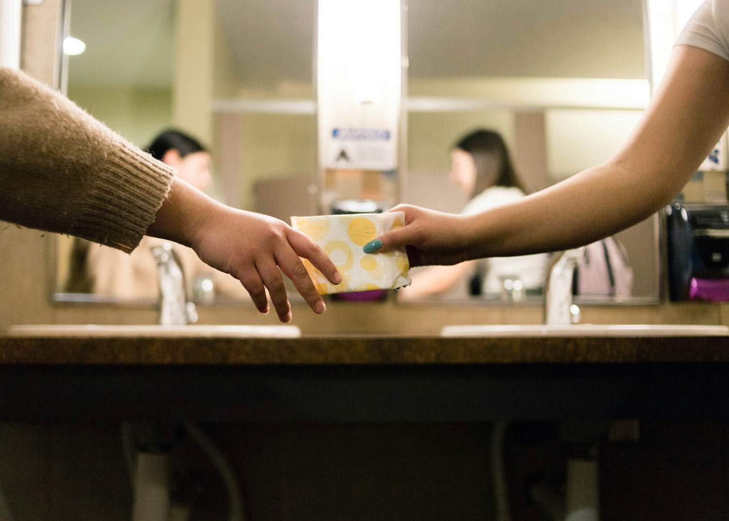 Two hands reaching towards each other across a bathroom counter, one holding a stack of paper towels with a patterned design.