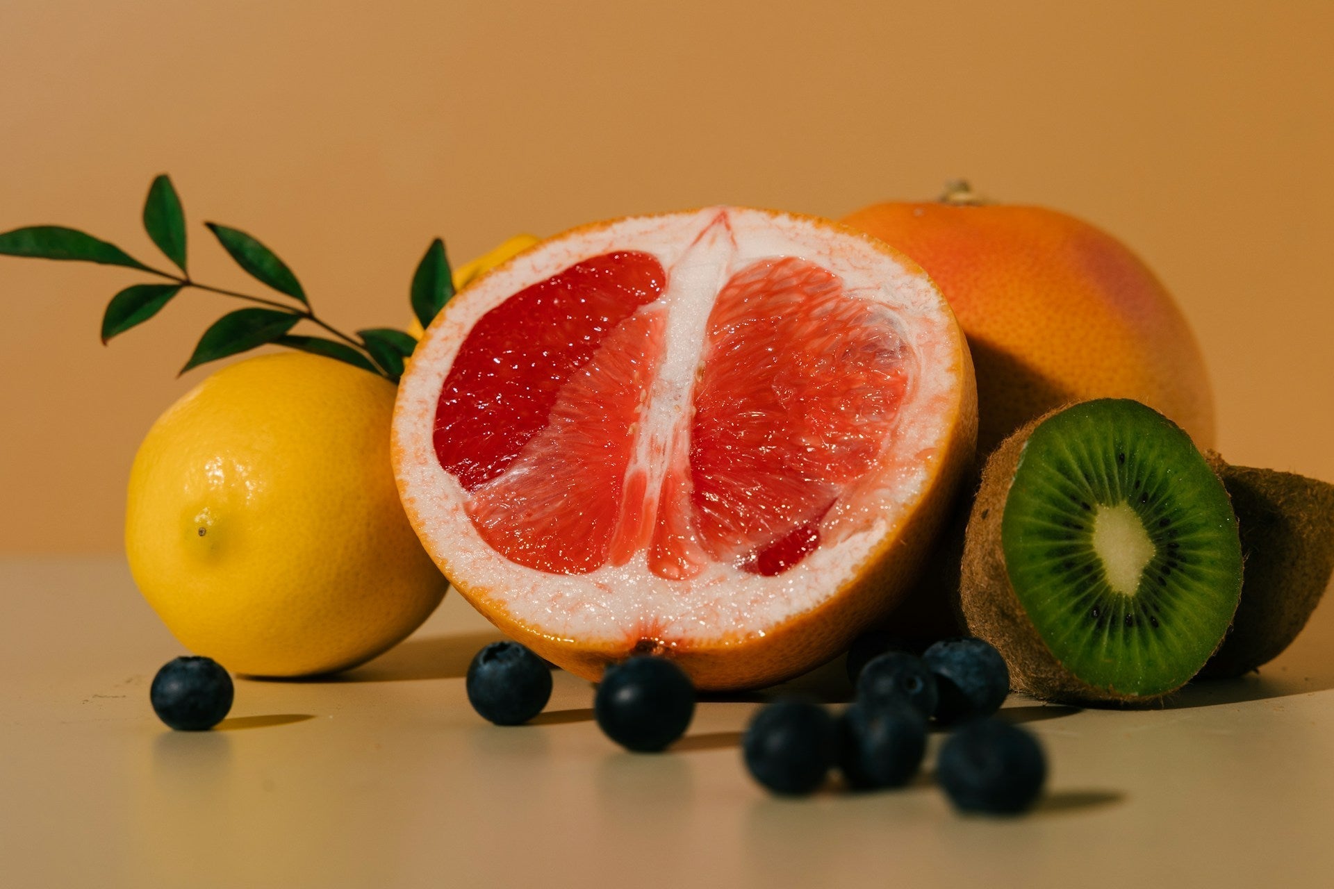 A vibrant display of fruits: a halved grapefruit, whole lemon, kiwi, and blueberries against a soft orange background.