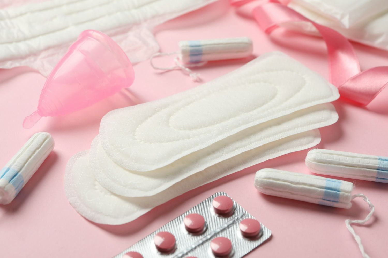 A collection of menstrual products including pads, tampons, a menstrual cup, pink pills, and pink ribbon on a pastel pink background.