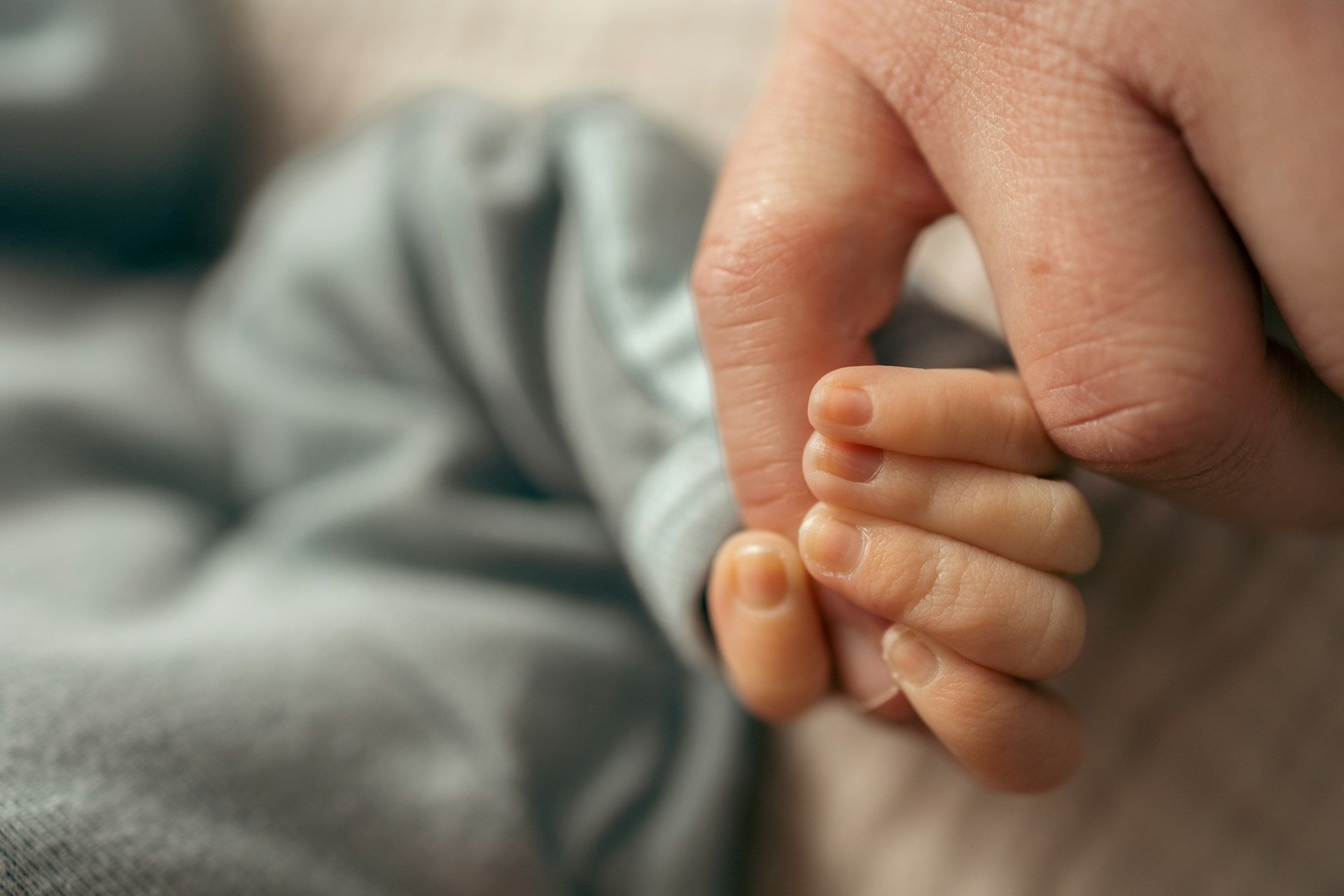Best postpartum pads: Close-up of an adult hand gently holding a newborn baby's hand against a soft gray fabric background.