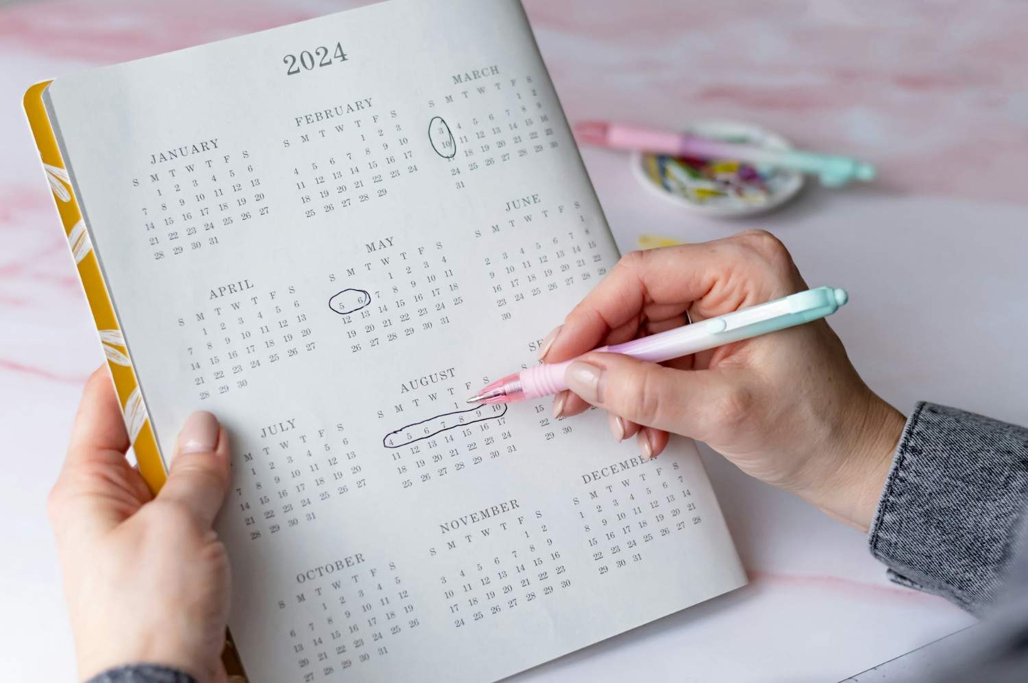 Find my flow period tool: A person writes in a 2024 calendar, marking dates in August and other months with colorful pens on a pink surface.