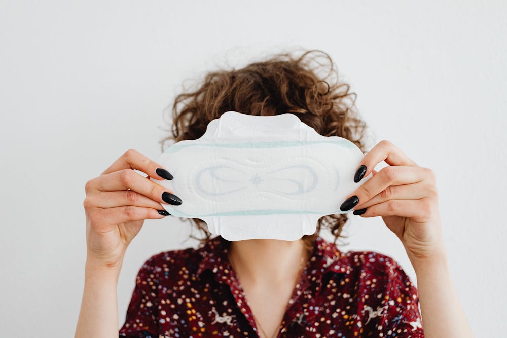 Fizabelle box contents: Person with curly hair and black nails holding a sanitary pad in front of their face against a white background.