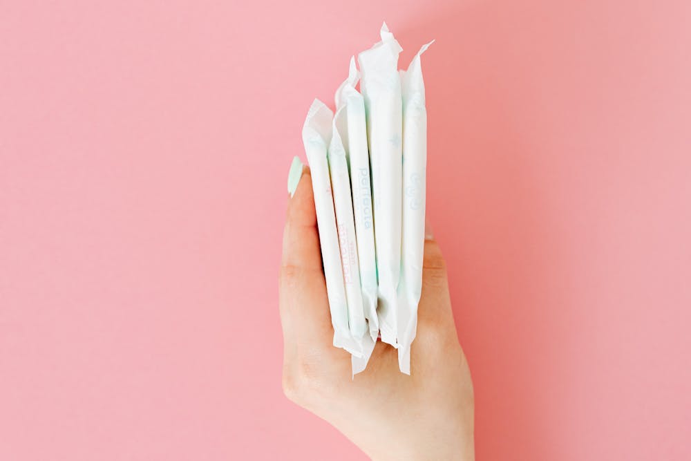 Adjust period subscription: Hand holding a stack of five individually wrapped white sanitary pads against a pink background.