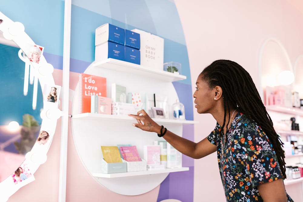 Curated period box: Person with long hair wearing a floral shirt selecting skincare or beauty products from a white circular wall shelf in a brightly colored store.