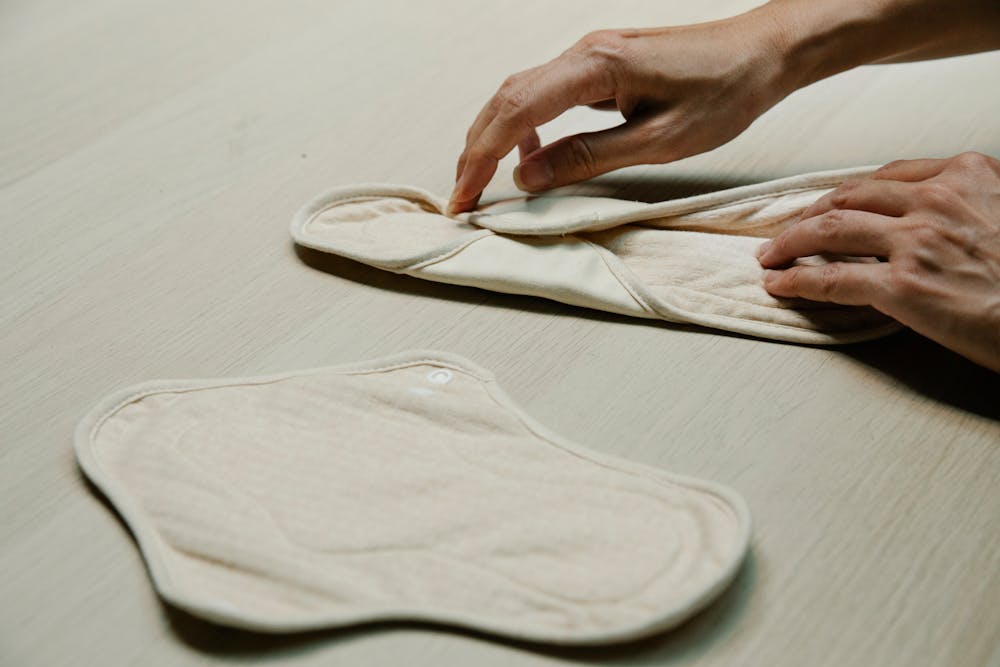 Custom period routine: A person’s hands are folding a soft, reusable cloth pad on a wooden surface, with another pad placed nearby.