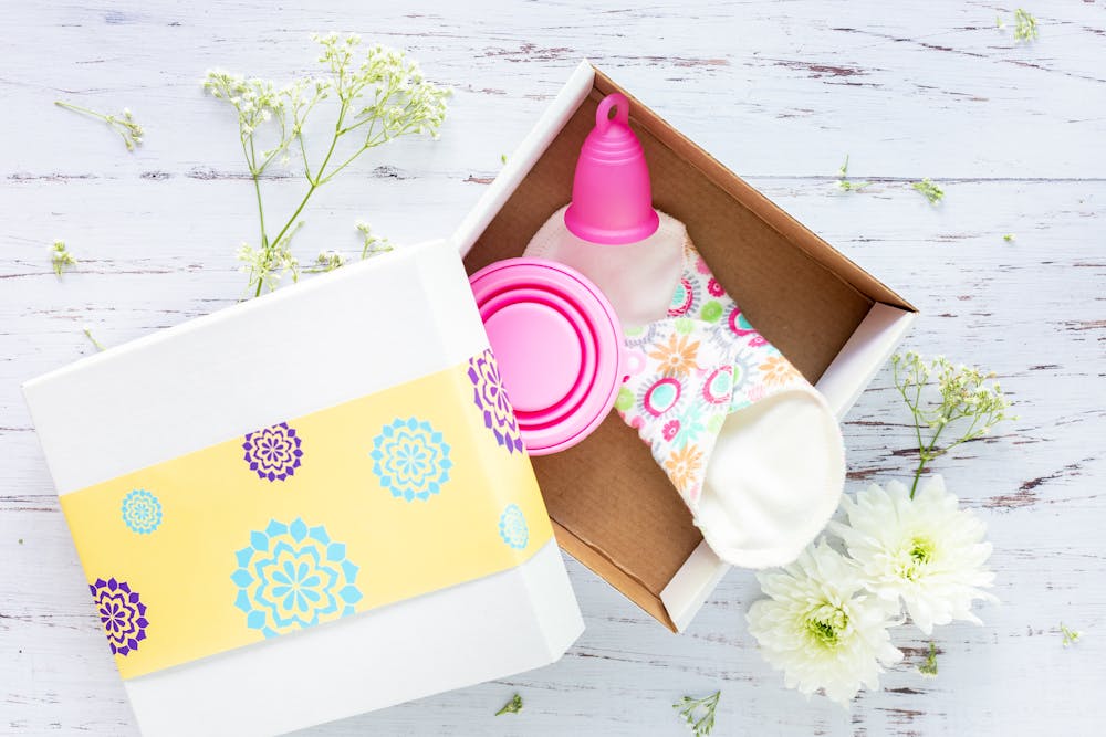 Period subscription box: Gift box with a pink menstrual cup, a collapsible cup, and a colorful floral cloth on a white wooden surface with white flowers.
